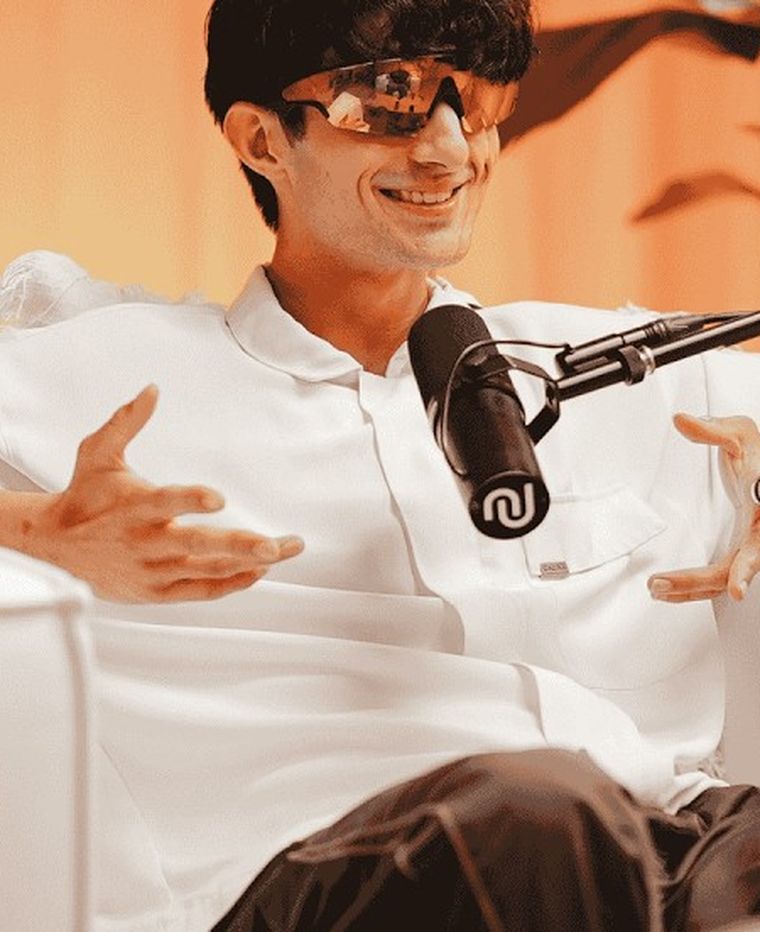 Matthew Byrden mid-conversation on a podcast set, seated on a white couch in front of a warm orange curtain.